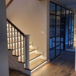 MEBLE Z PASJĄ Emilia Obłąk - Nowoczesne schody z podświetleniem LED na stopniach i balustradzie, drewniane belki stropowe, podłoga z paneli w odcieniach brązu, przeszklona ściana działowa z czarnymi szprosami.