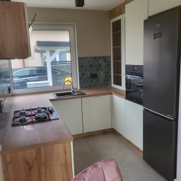 MEBLE Z PASJĄ Emilia Obłąk - Nowoczesna kuchnia z drewnianymi blatami i białymi szafkami, heksagonalne płytki nad blatem, czarna płyta gazowa, lodówka w kolorze grafitowym i krzesło z różowego weluru.