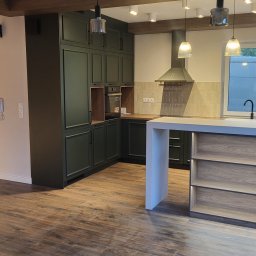 MEBLE Z PASJĄ Emilia Obłąk - Nowoczesna kuchnia z ciemnozielonymi szafkami, drewnianym blatem i wyspą z jasnego drewna, oświetlona wiszącymi lampami z szarymi kloszami, z podłogą wyłożoną panelami imitującymi drewno.