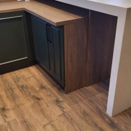 MEBLE Z PASJĄ Emilia Obłąk - Nowoczesna kuchnia z ciemnozielonymi szafkami, blatem imitującym drewno i białym półwyspem, widoczna na tle okna z zielenią za nim.