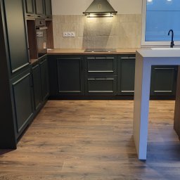 MEBLE Z PASJĄ Emilia Obłąk - Nowoczesna kuchnia z ciemnozielonymi szafkami, drewnianym blatem i brązowymi panelami podłogowymi imitującymi drewno. Widoczny okap i okno z widokiem na zewnątrz. Oświetlenie sufitowe punktowe.