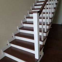 MEBLE Z PASJĄ Emilia Obłąk - Nowoczesne schody z ciemnego drewna z białymi balustradami i listwami przypodłogowymi, widok z dołu, z widocznymi otworami po montażu oświetlenia.