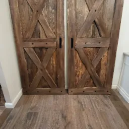 Dwie przesuwne drzwi stodołowe z ciemnego drewna, z czarnymi uchwytami, zamontowane wewnątrz pomieszczenia z podłogą z paneli imitujących drewno, widoczne z niskiego kąta.
