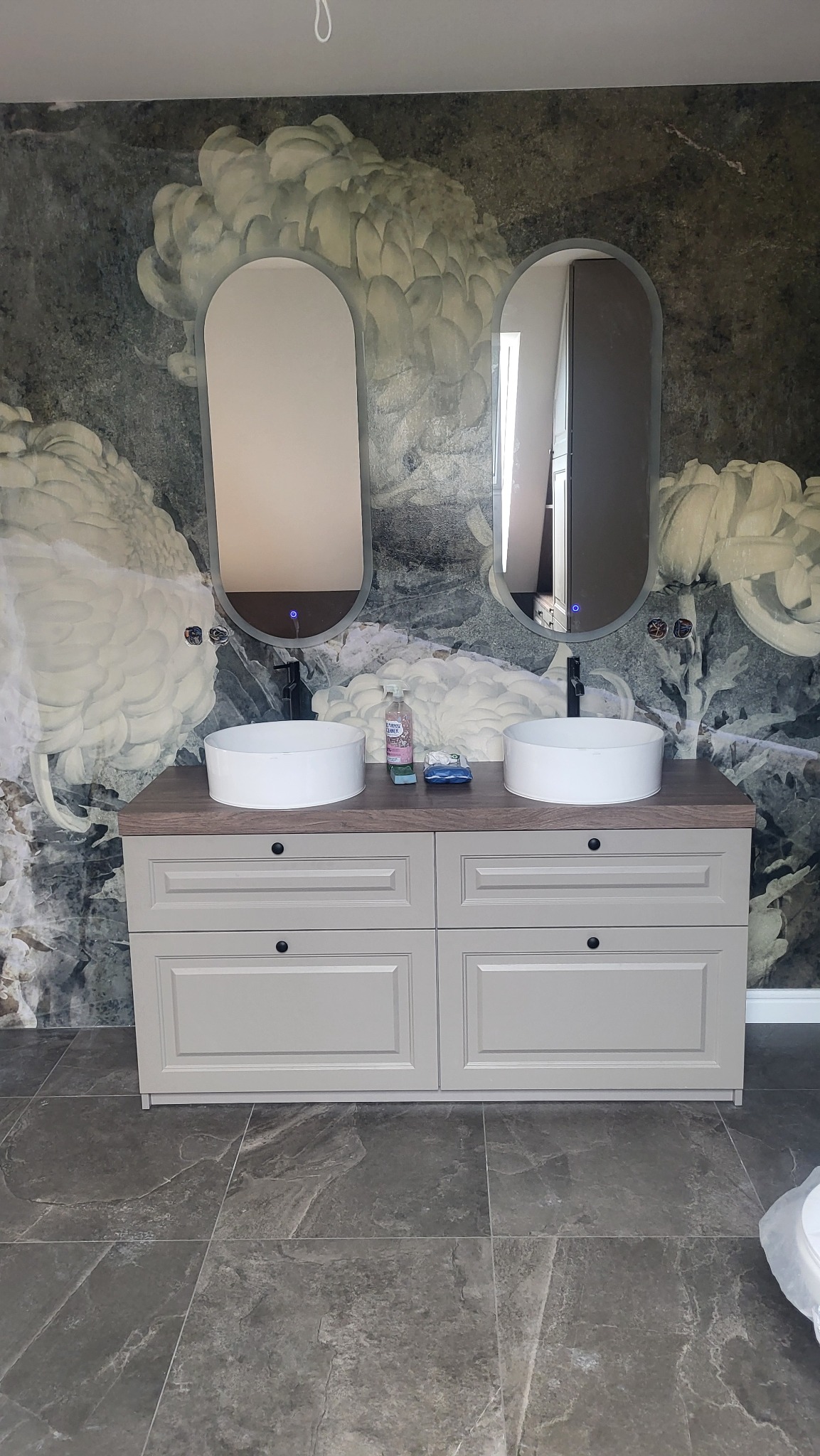 Łazienka z dwoma umywalkami nablatowymi, szafką w stylu retro z szufladami i dwoma owalnymi lustrami z podświetleniem LED na tle ściany z tapetą w kwiaty.