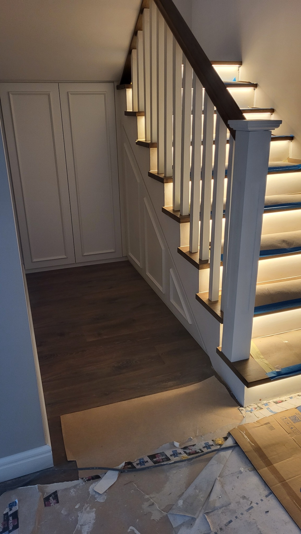 Podświetlane schody z białymi balustradami i drewnianymi stopniami, widoczne zabezpieczenia na stopniach i podłoga z panelami, w tle białe szafy w zabudowie.