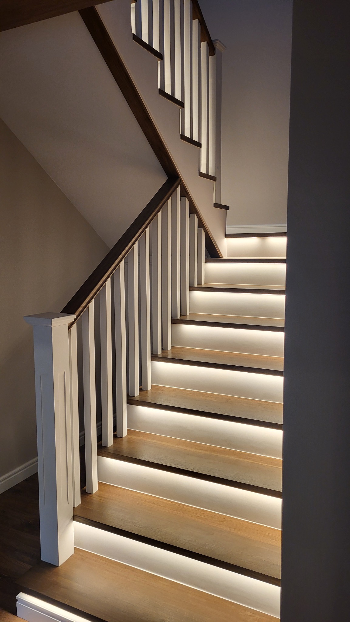 Drewniane schody z białymi balustradami i podświetleniem LED na każdym stopniu, tworzącym ciepłą, nowoczesną atmosferę.