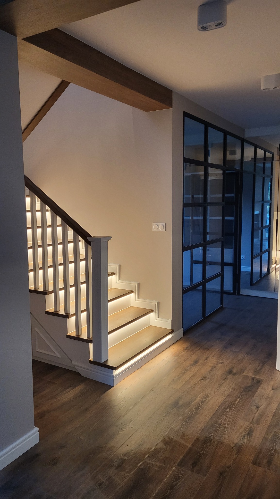 Nowoczesne schody z podświetleniem LED na stopniach i balustradzie, drewniane belki stropowe, podłoga z paneli w odcieniach brązu, przeszklona ściana działowa z czarnymi szprosami.