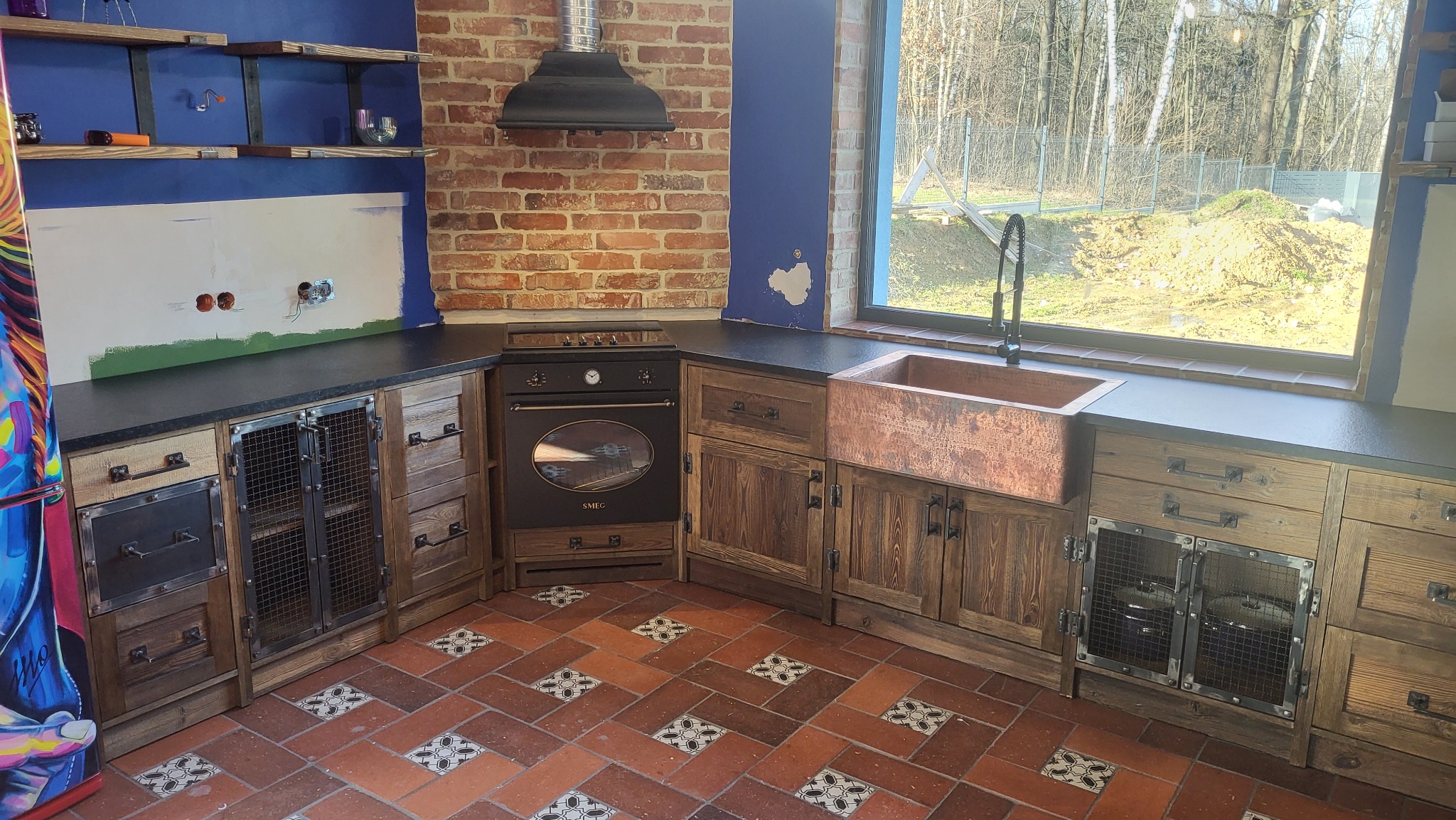 Rustykalna kuchnia z drewnianymi szafkami z metalowymi siatkami, miedzianym zlewem, ceglaną ścianą i terakotową podłogą z dekoracyjnymi wstawkami. Ściana nad blatem częściowo przygotowana...