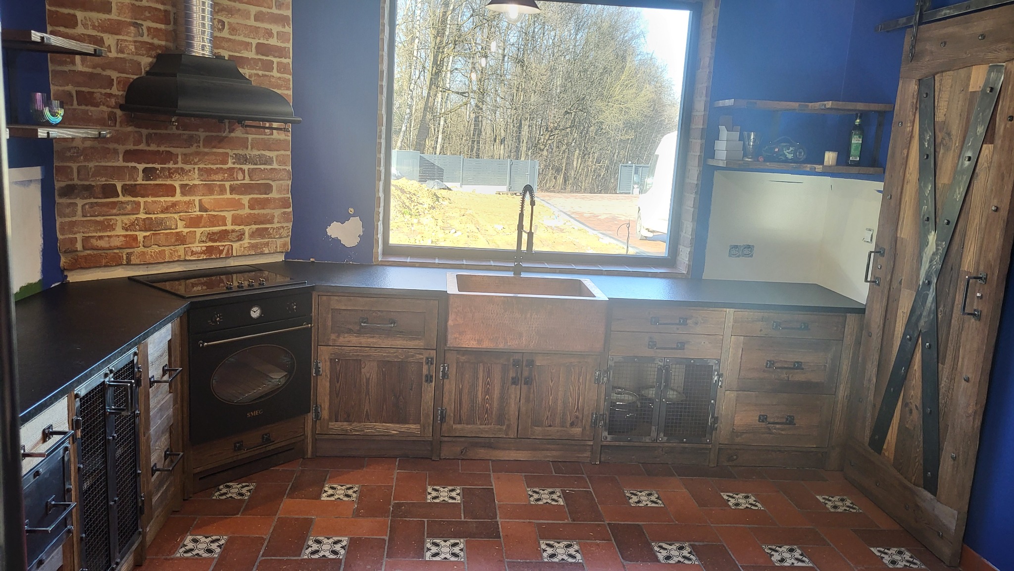 Rustykalna kuchnia z miedzianym zlewem, drewnianymi szafkami z metalowymi kratkami, ceglaną ścianą z okapem i przesuwnymi drzwiami stodołowymi, uzupełniona terakotową podłogą z dekoracyjnymi płytkami.