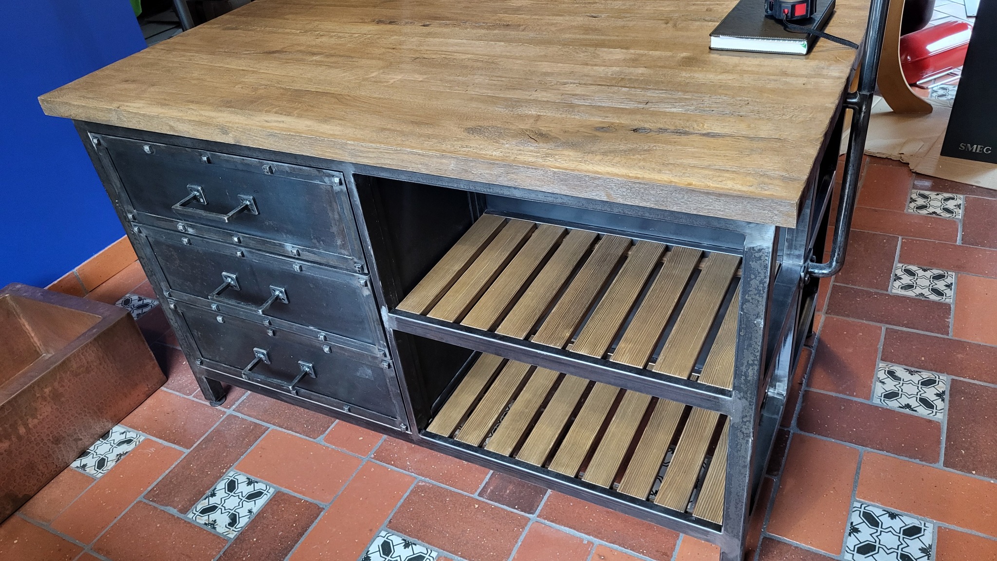 Metalowa szafka z drewnianym blatem i szufladami w stylu industrialnym, z drewnianymi półkami, stojąca na podłodze z terakoty z dekoracyjnymi wstawkami.
