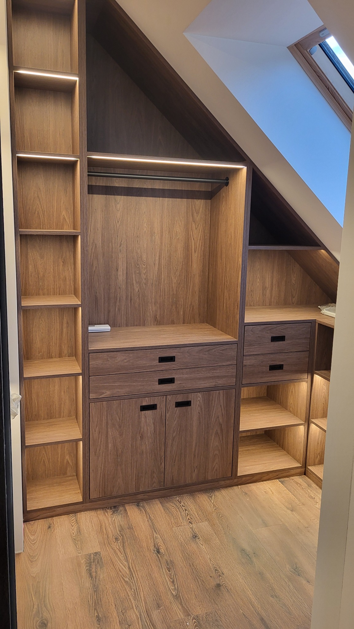 Zabudowa szafy wnękowej z drewna w pokoju na poddaszu, z oświetleniem LED w półkach i drążkiem na ubrania.