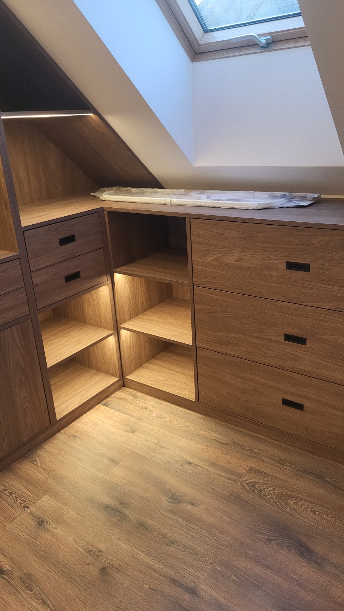 Zabudowa pod skosem z drewna z szufladami i otwartymi półkami z podświetleniem LED, widoczny fragment okna dachowego.
