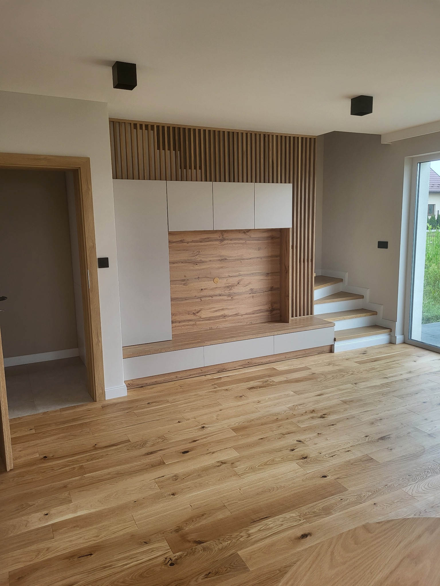 Nowoczesny salon z drewnianą podłogą, zabudową TV z drewnianymi lamelami i schodami z białymi stopnicami, oświetlony czarnymi lampami sufitowymi.