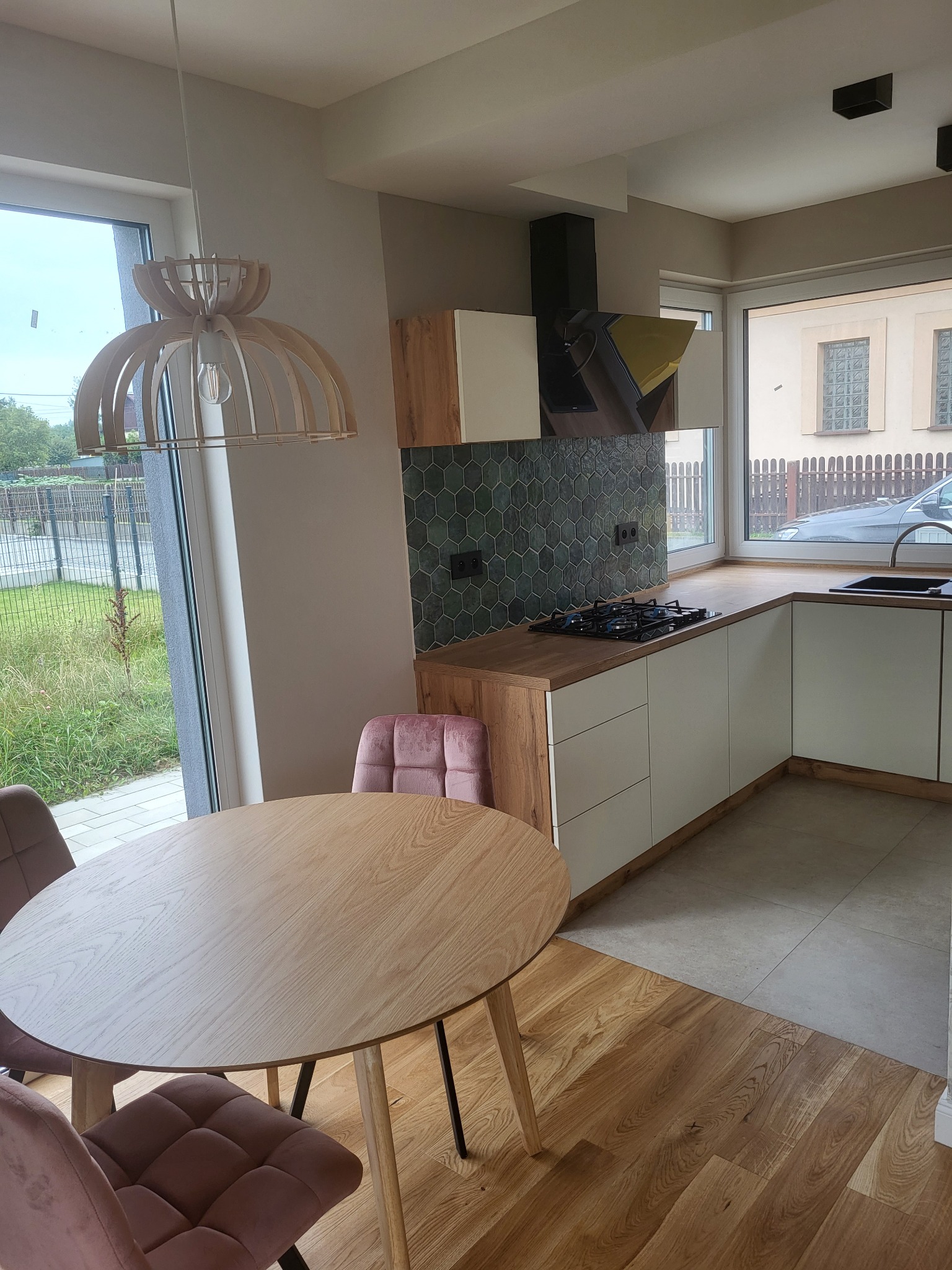 Jasna kuchnia z drewnianym stołem i krzesłami, wisząca drewniana lampa, heksagonalne płytki na ścianie, widok na ogród za oknem.