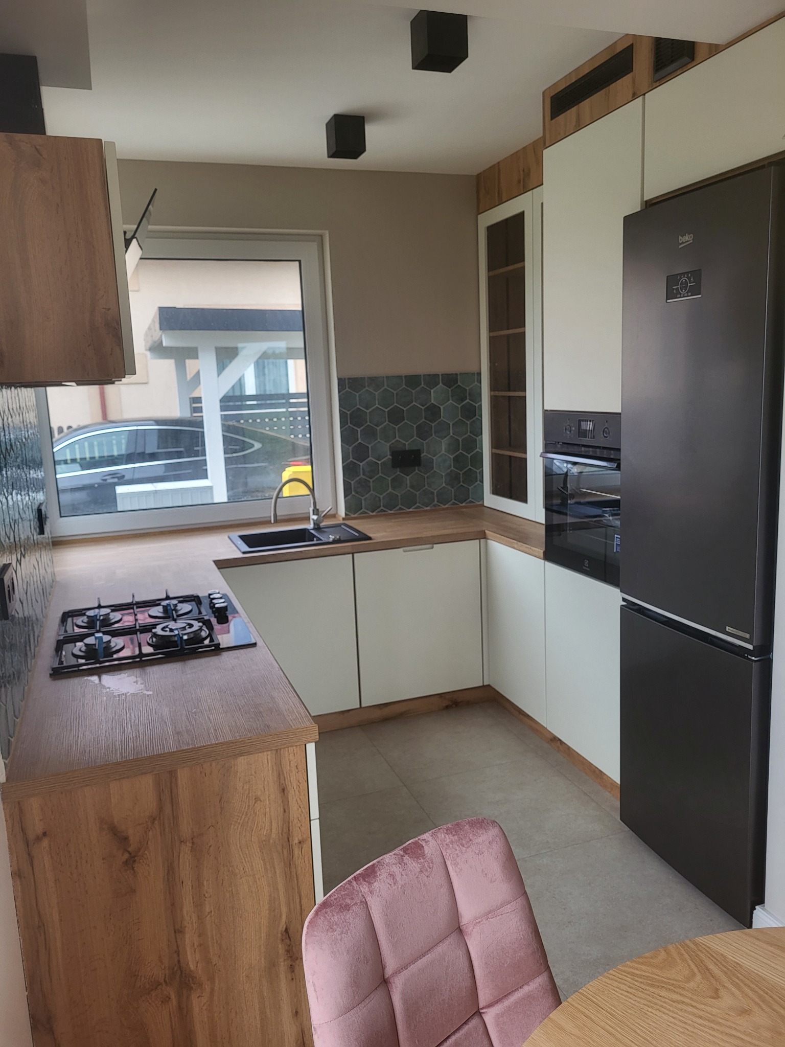 Nowoczesna kuchnia z drewnianymi blatami i białymi szafkami, heksagonalne płytki nad blatem, czarna płyta gazowa, lodówka w kolorze grafitowym i krzesło z różowego weluru.