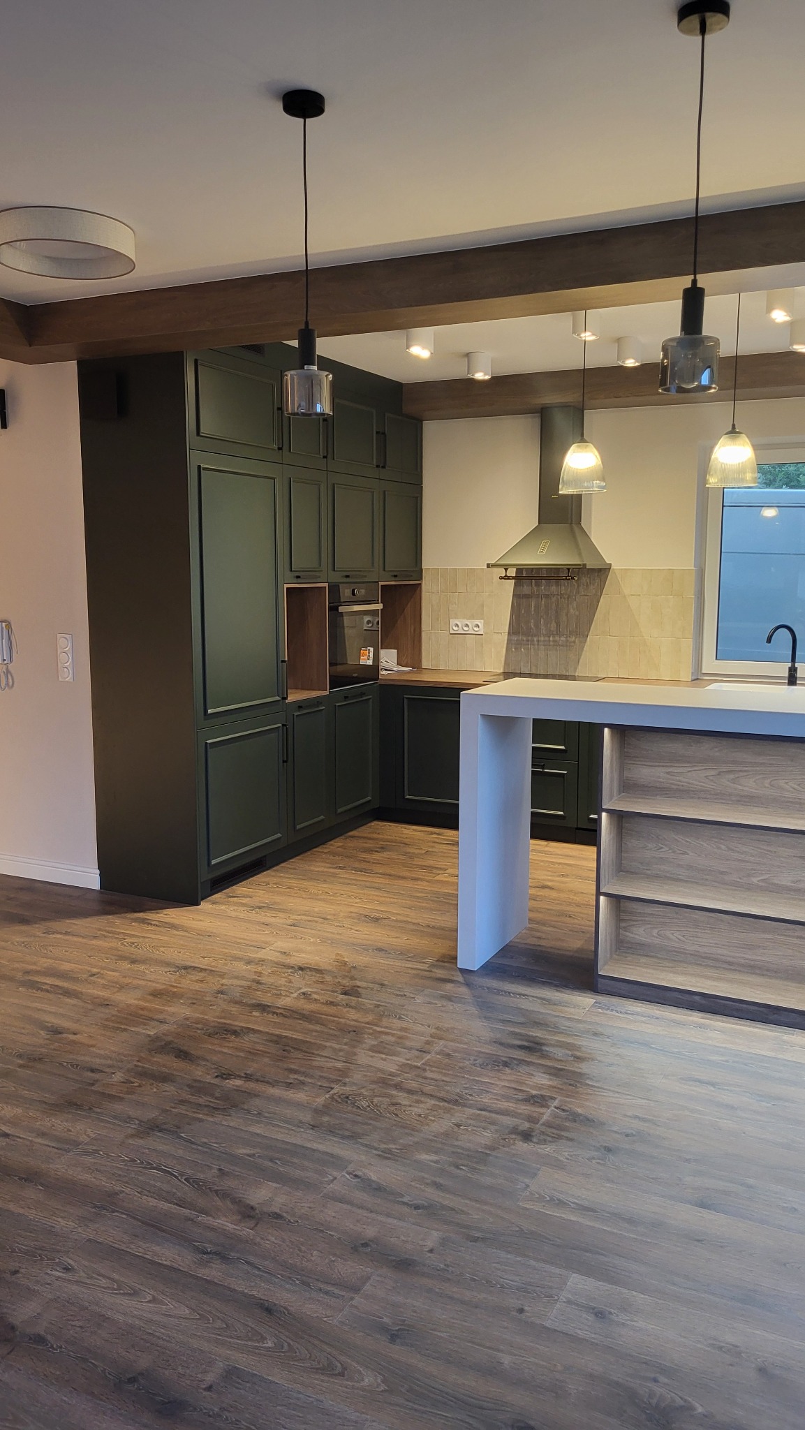 Nowoczesna kuchnia z ciemnozielonymi szafkami, drewnianym blatem i wyspą z jasnego drewna, oświetlona wiszącymi lampami z szarymi kloszami, z podłogą wyłożoną panelami imitującymi drewno.
