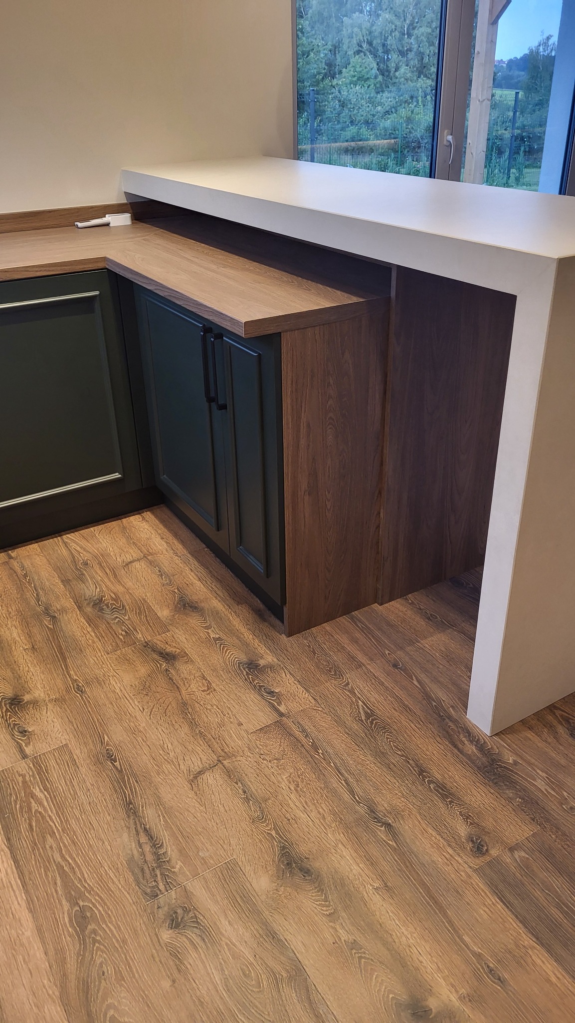 Nowoczesna kuchnia z ciemnozielonymi szafkami, blatem imitującym drewno i białym półwyspem, widoczna na tle okna z zielenią za nim.