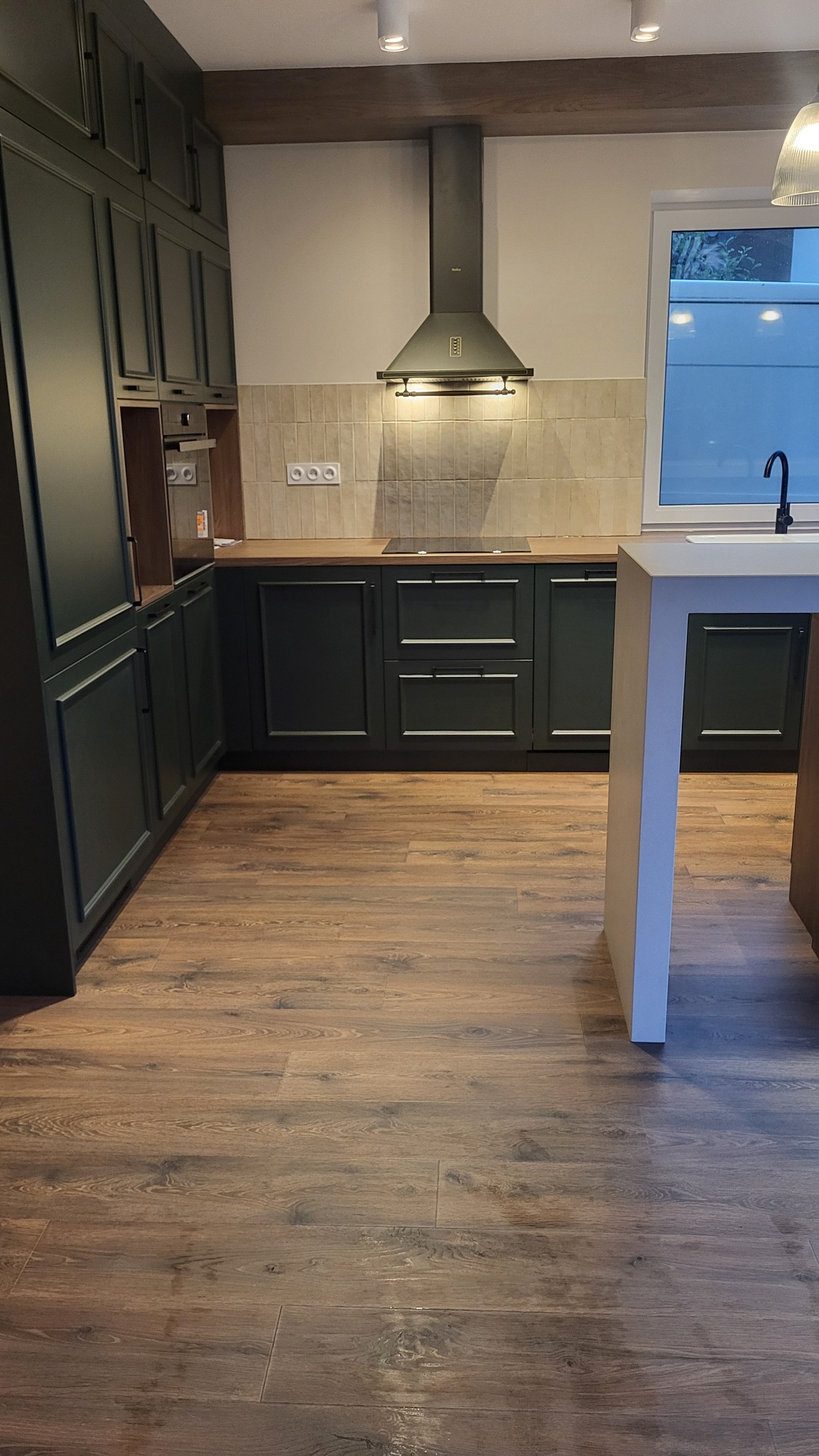 Nowoczesna kuchnia z ciemnozielonymi szafkami, drewnianym blatem i brązowymi panelami podłogowymi imitującymi drewno. Widoczny okap i okno z widokiem na zewnątrz. Oświetlenie sufitowe punktowe.