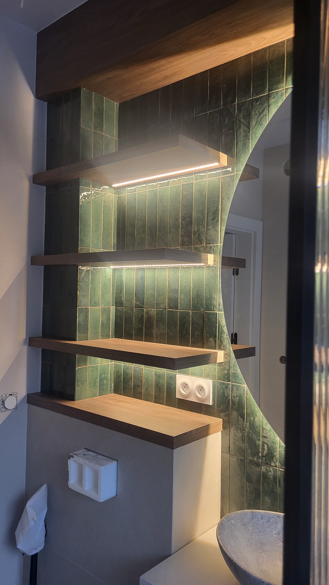 Nowoczesna łazienka z zielonymi, pionowymi płytkami, drewnianymi półkami podświetlonymi LED, okrągłym lustrem z podświetleniem i umywalką z kamienia.