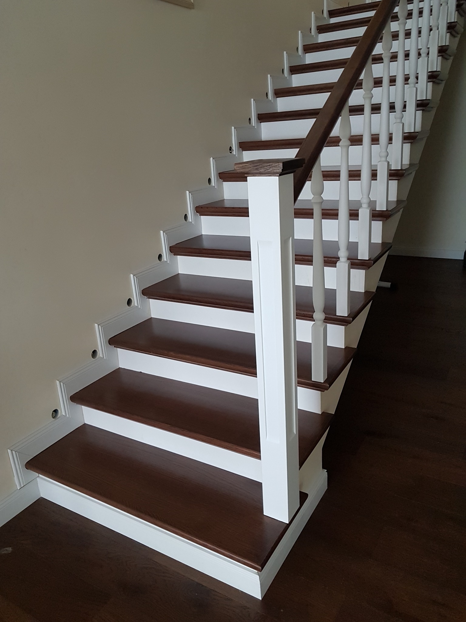 Nowoczesne schody z ciemnego drewna z białymi balustradami i listwami przypodłogowymi, widok z dołu, z widocznymi otworami po montażu oświetlenia.