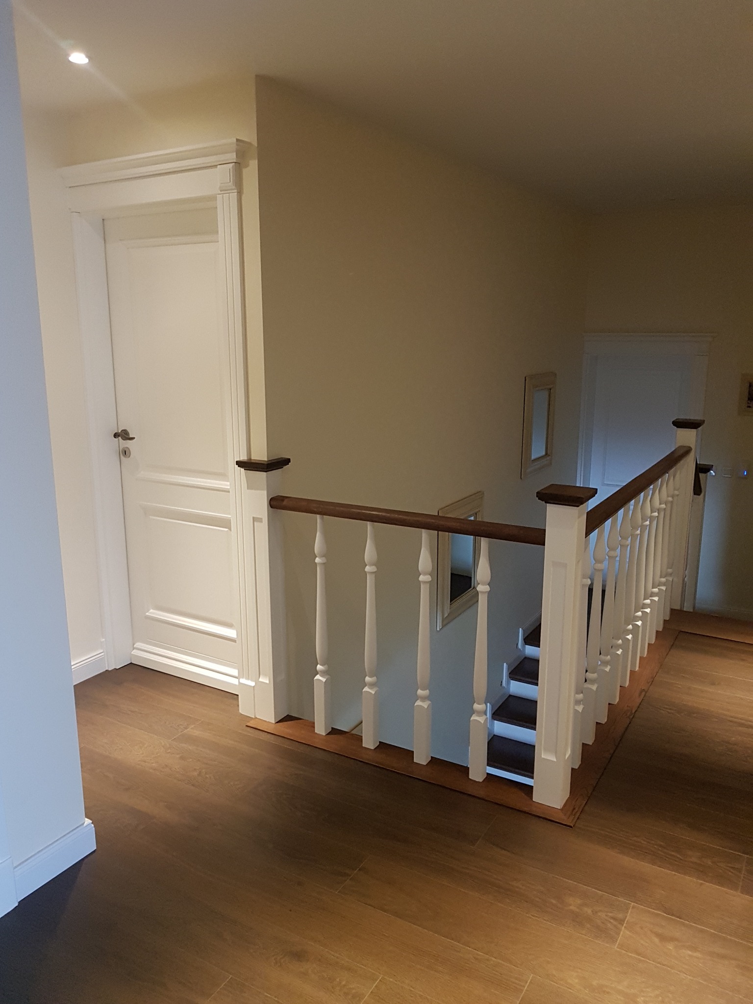 Korytarz z brązowymi panelami podłogowymi, białe drzwi z ozdobną ramą, schody z białą balustradą i drewnianym poręczą, widoczne fragmenty luster na ścianie.