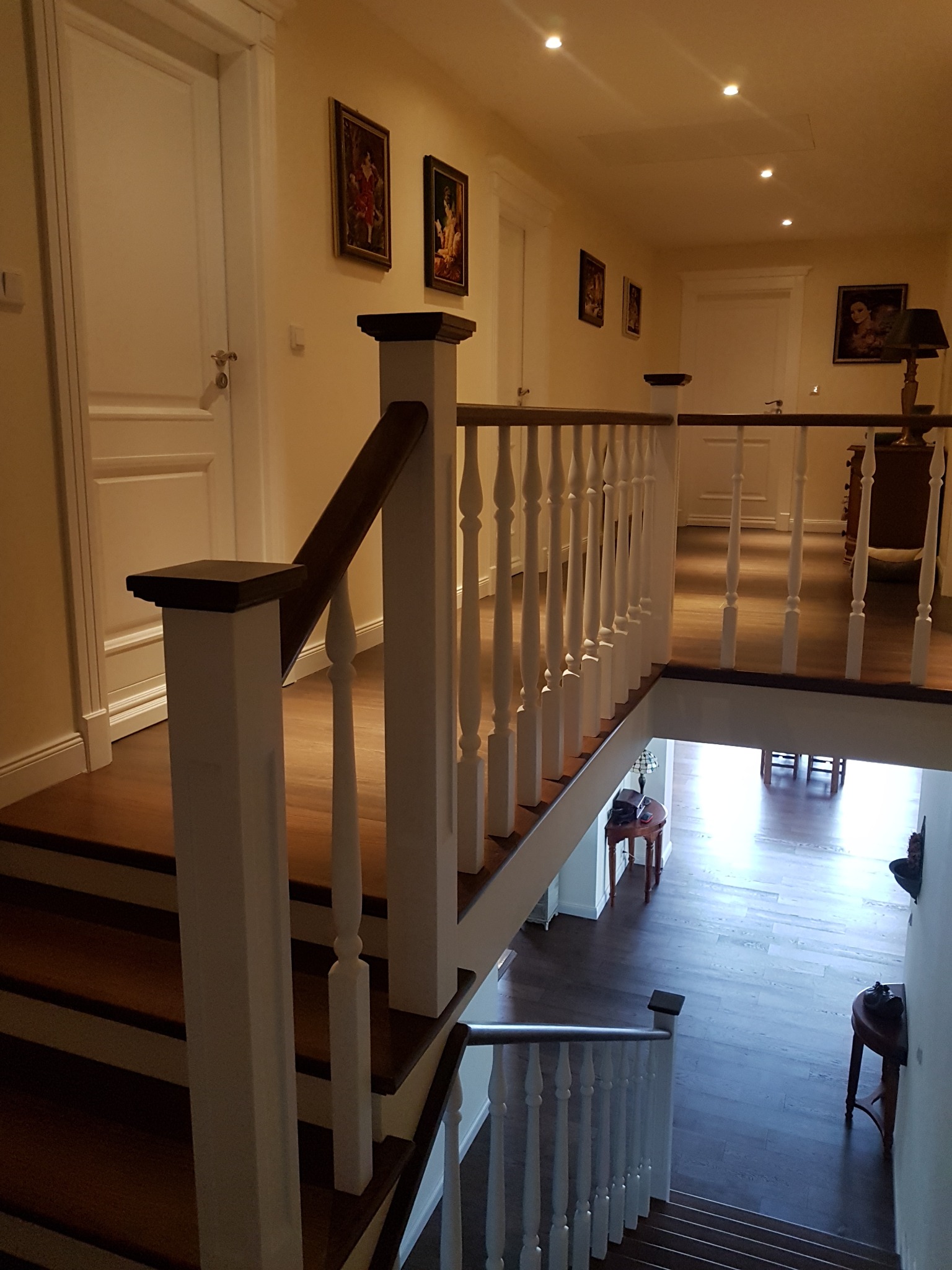 Klasyczne schody drewniane z białymi balustradami i ciemnobrązowym poręczą, widok z góry na korytarz z obrazami na ścianach i jasnymi drzwiami.