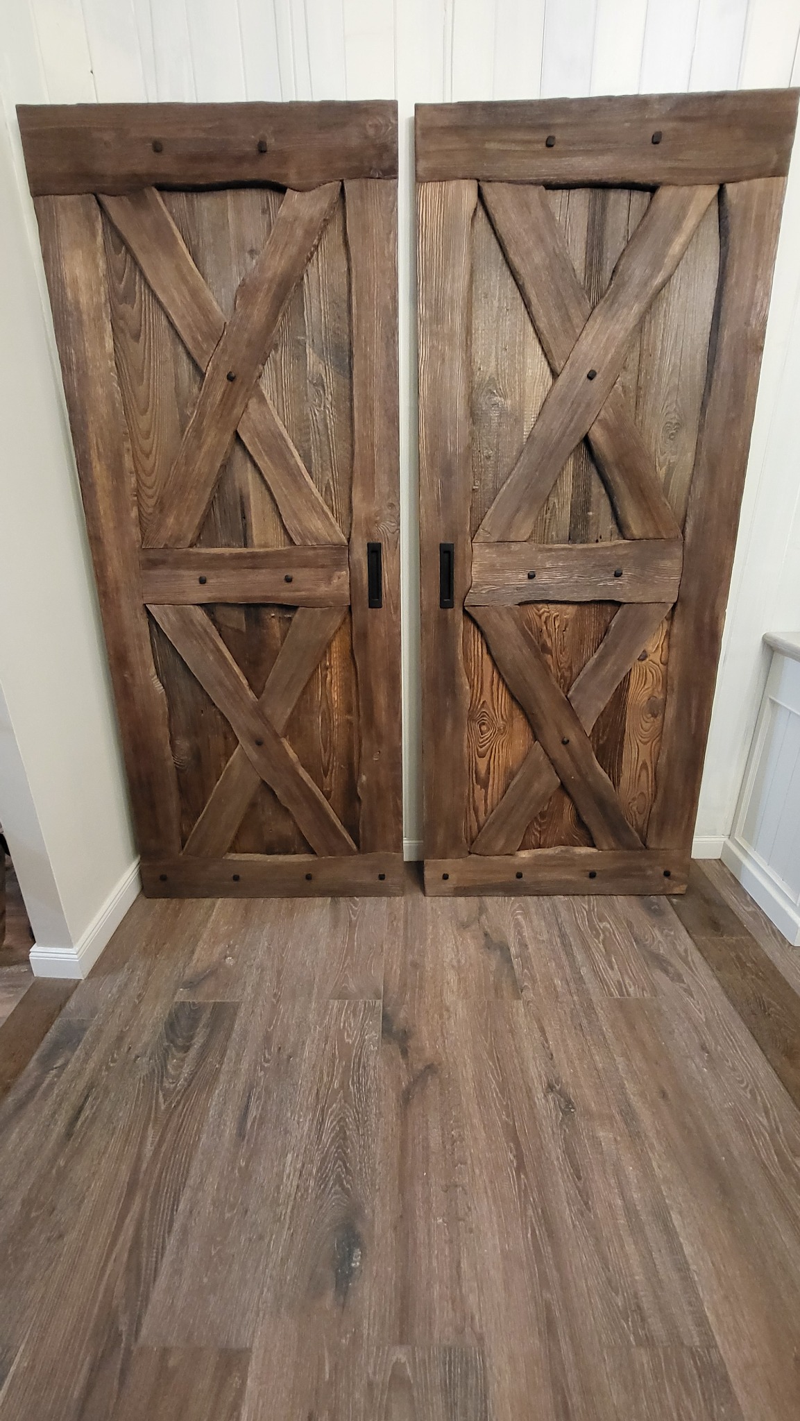 Dwie przesuwne drzwi stodołowe z ciemnego drewna, z czarnymi uchwytami, zamontowane wewnątrz pomieszczenia z podłogą z paneli imitujących drewno, widoczne z niskiego kąta.