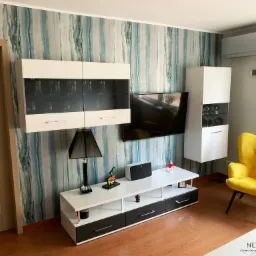 Nowoczesny salon z akcentem w postaci tapety w odcieniach błękitu i beżu, wiszące szafki z przeszklonymi frontami, biała szafka RTV, żółty fotel typu muszelka i klimatyzator na szarej ścianie.