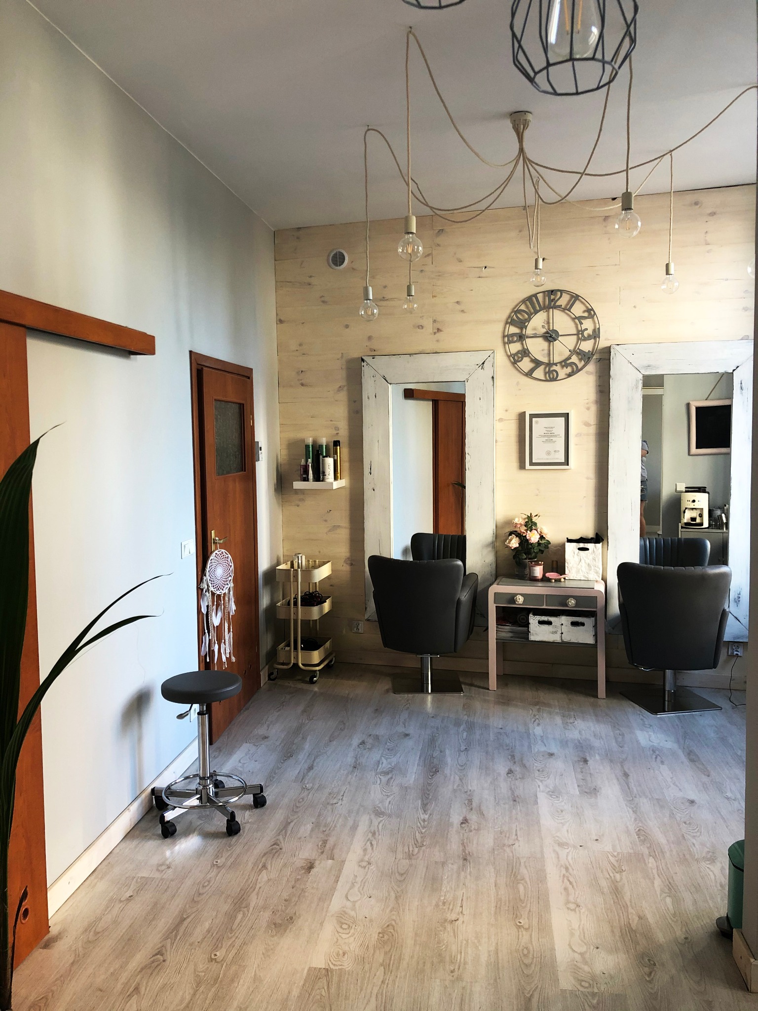 Jasne wnętrze salonu z drewnianymi panelami ściennymi, dwoma stanowiskami z lustrami w białych ramach, krzesłami fryzjerskimi, dekoracyjnym zegarem na ścianie i oryginalnym oświetleniem zwisającym...