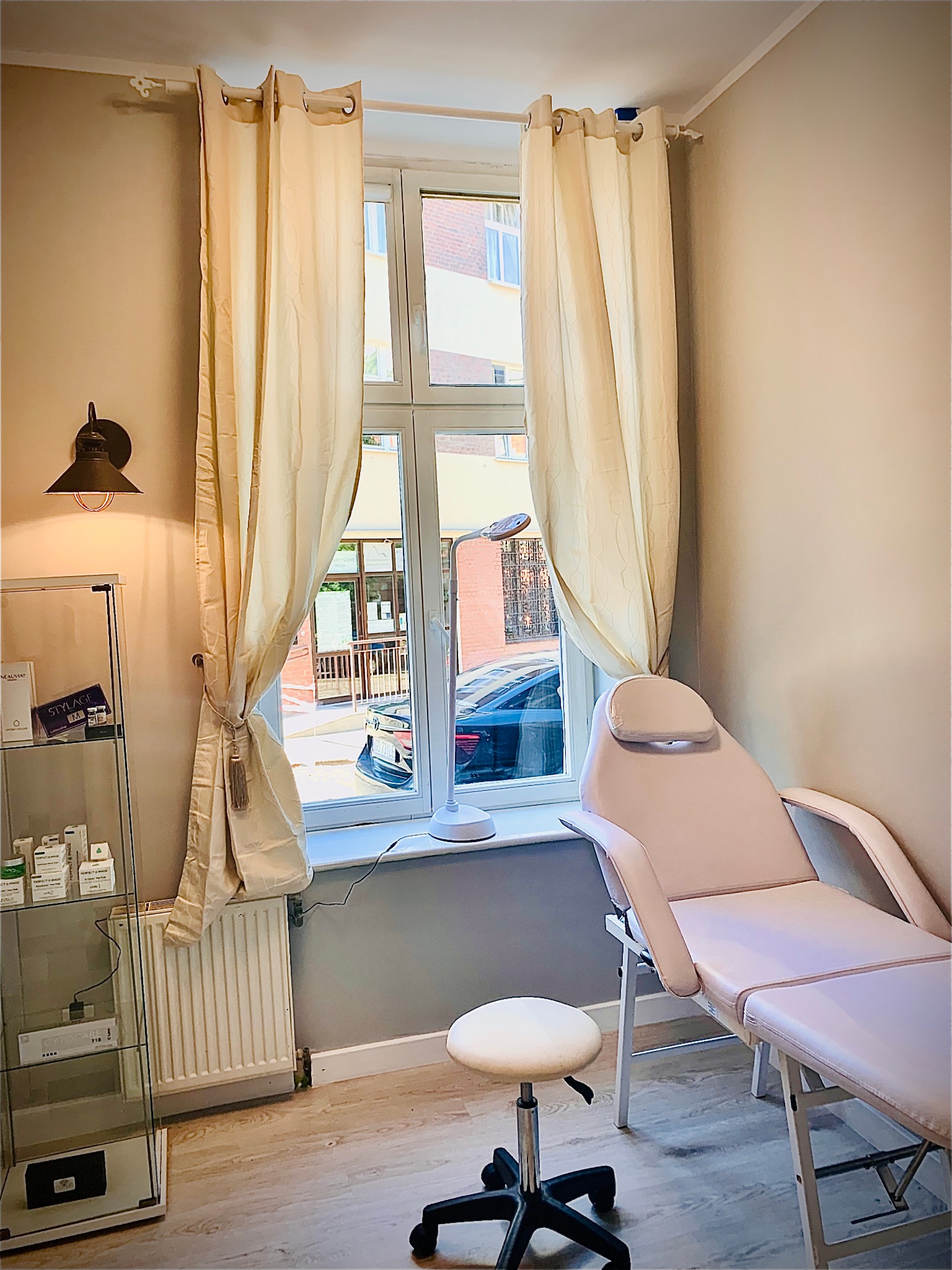 Jasne wnętrze gabinetu kosmetycznego z fotelem zabiegowym, okrągłym stołkiem na kółkach i szklaną witryną z kosmetykami, oświetlone lampą ścienną w stylu retro oraz naturalnym światłem z okna...