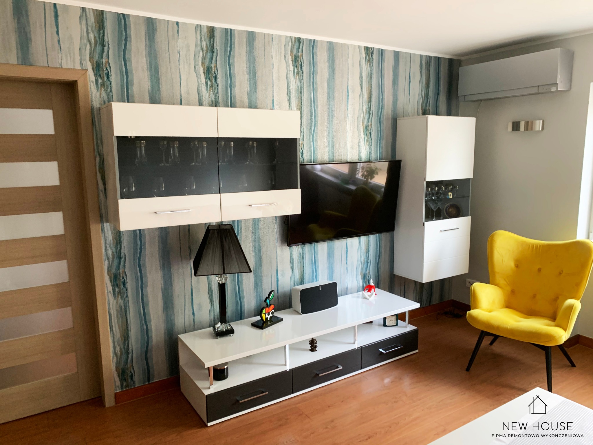 Nowoczesny salon z akcentem w postaci tapety w odcieniach błękitu i beżu, wiszące szafki z przeszklonymi frontami, biała szafka RTV, żółty fotel typu muszelka i klimatyzator na szarej ścianie.