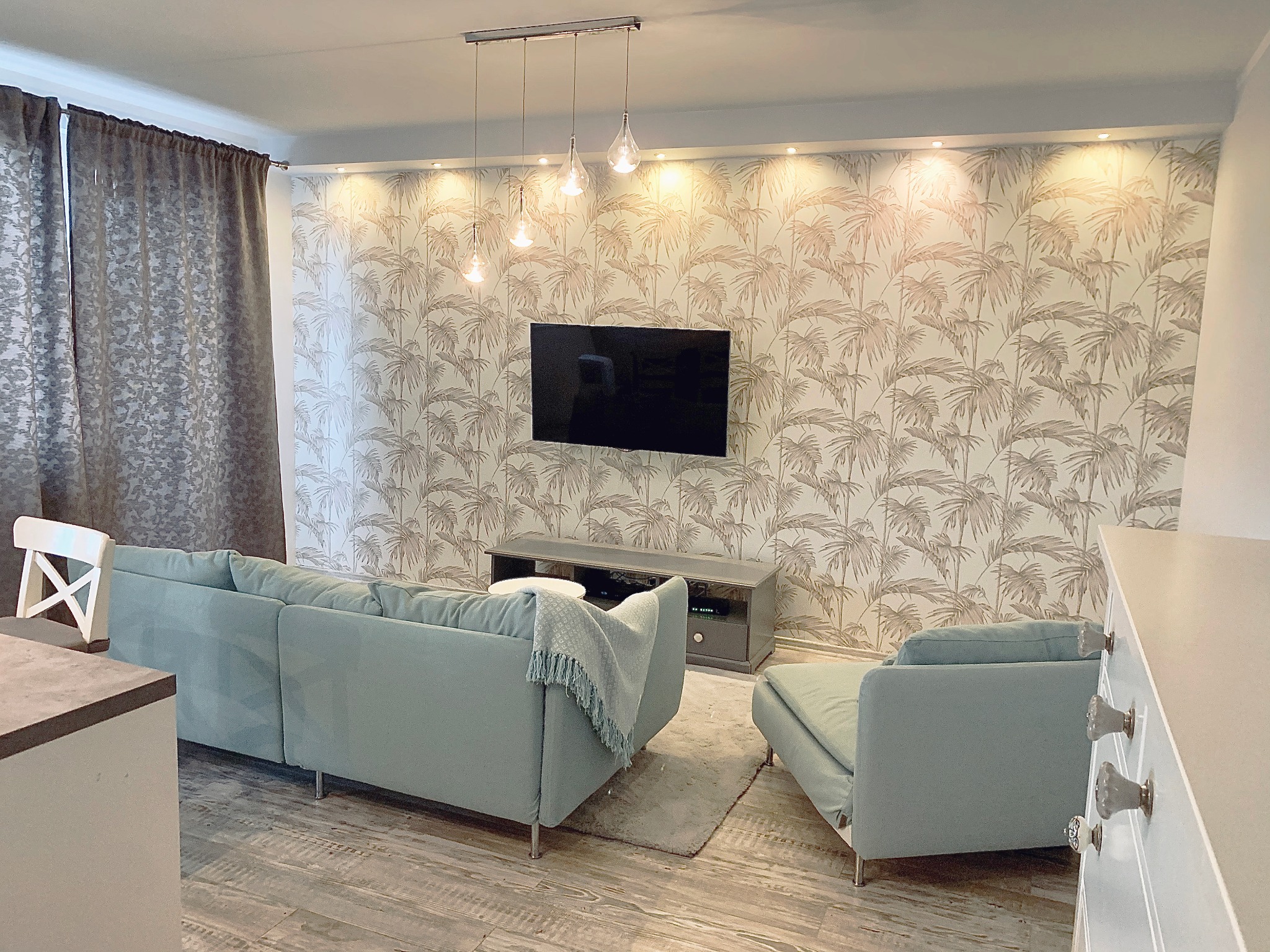 Jasny salon z turkusową sofą i fotelem, drewnopodobna podłoga, ściana w tapetę z motywem palm, oświetlenie punktowe i wiszące lampy. Minimalistyczny wystrój.