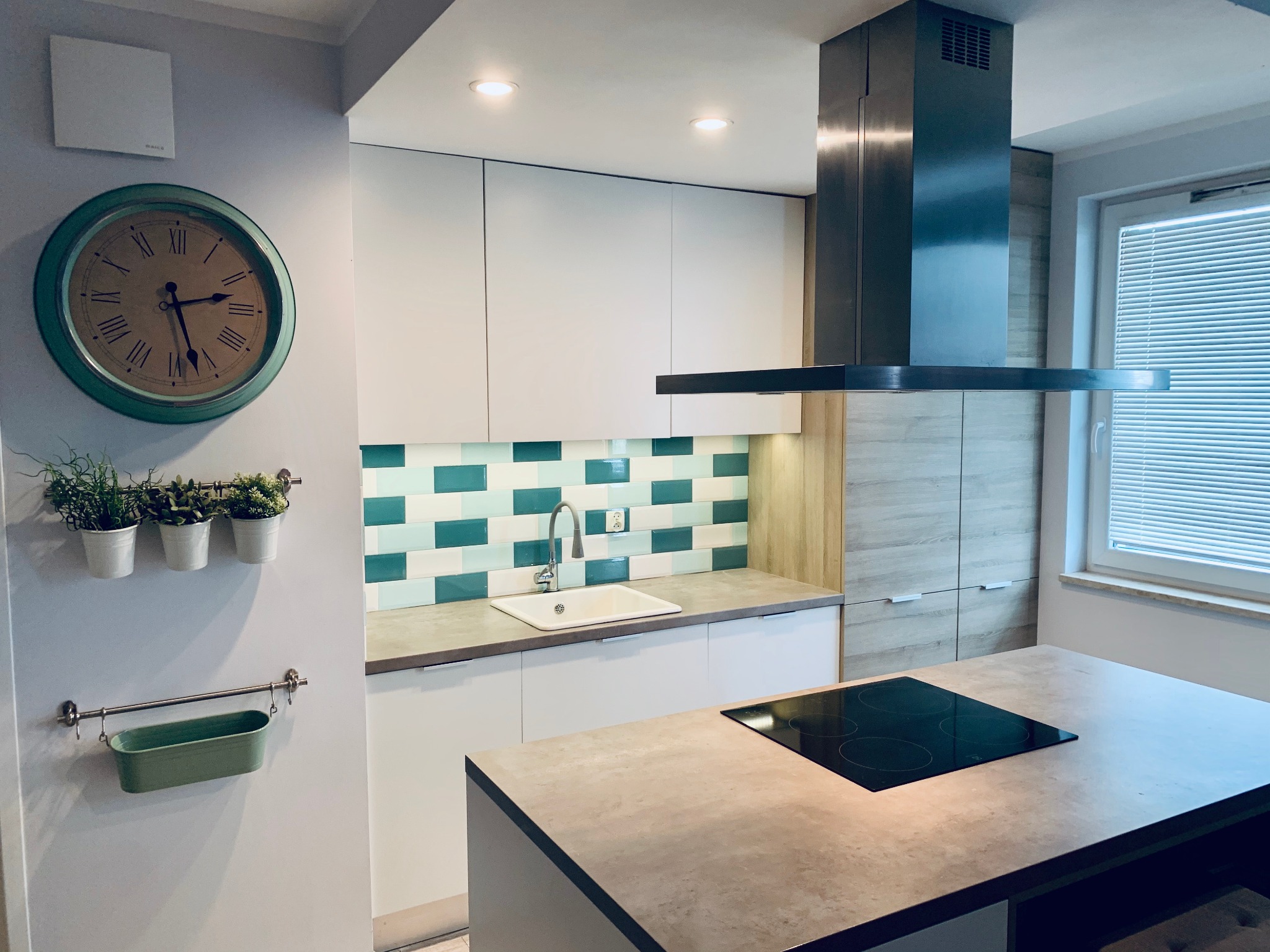 Nowoczesna kuchnia z białymi szafkami, wyspą z płytą indukcyjną, zielono-białymi płytkami nad blatem, wiszącymi ziołami i dużym zegarem w stylu retro.