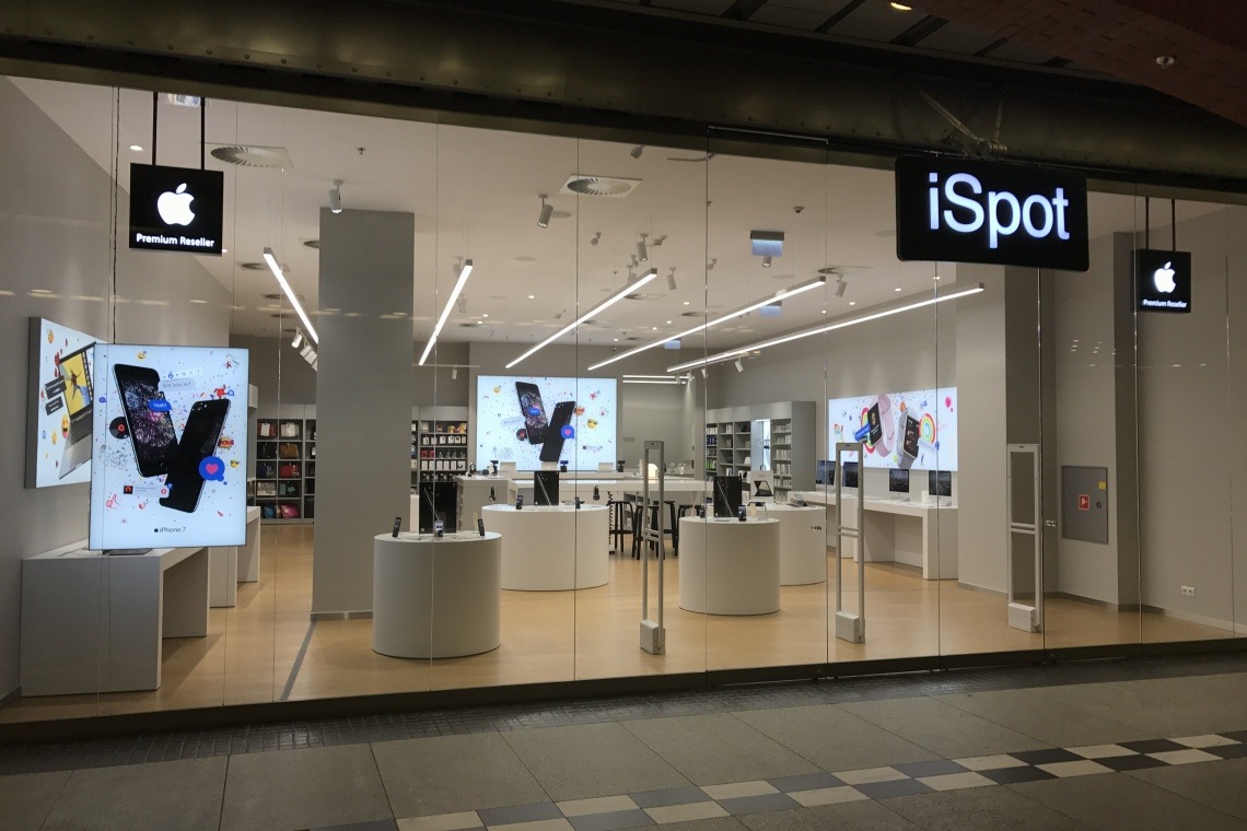 Salon iSpot w galerii handlowej Stary Browar w Poznaniu, widok z zewnątrz przez szklaną witrynę. Długie, liniowe lampy sufitowe oświetlają ekspozycję urządzeń Apple.