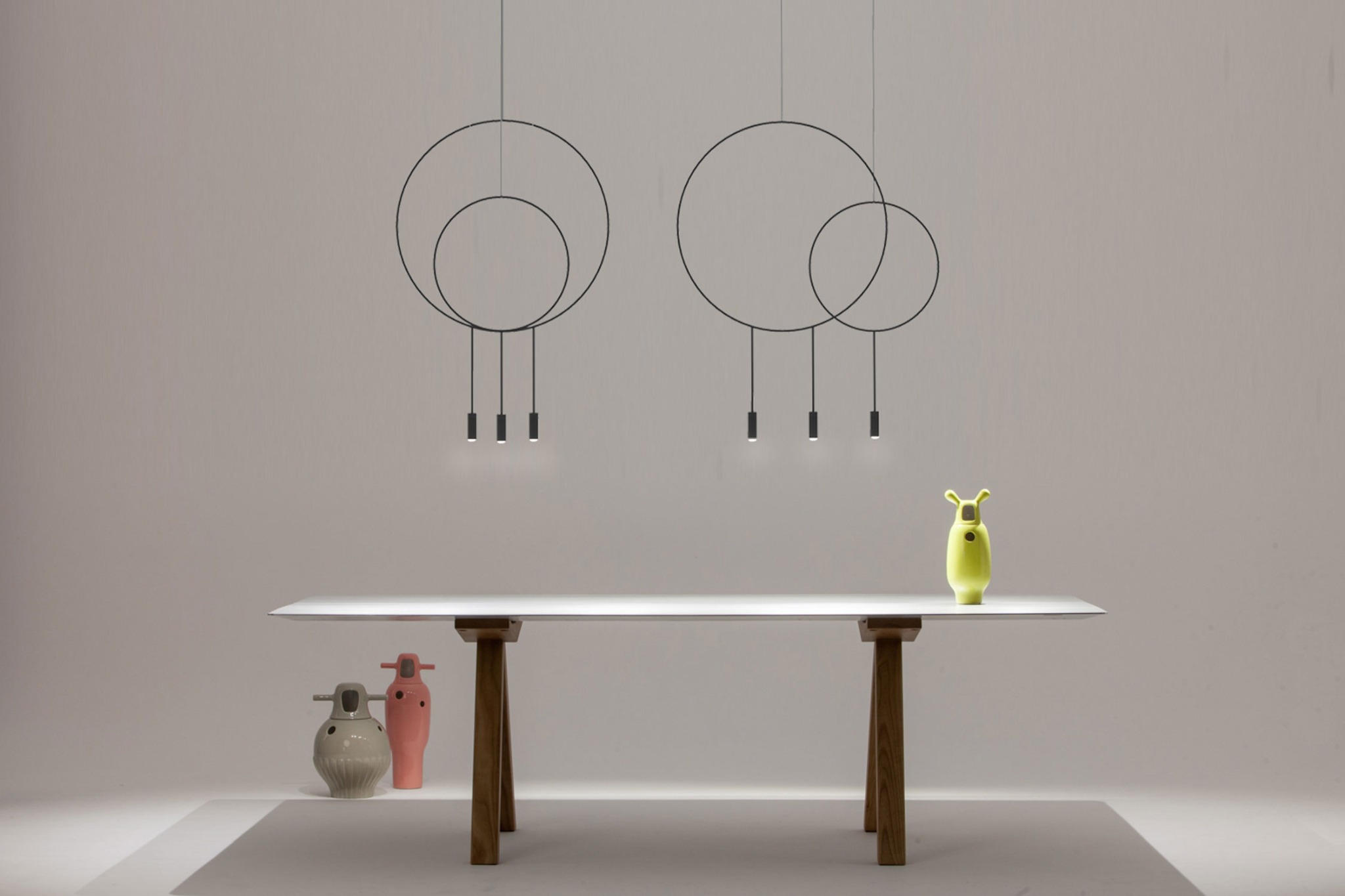 Dwa nowoczesne, okrągłe żyrandole z czarnymi obwodami i trzema punktami świetlnymi każdy, zawieszone nad białym stołem z drewnianymi nogami. Na stole stoi zielona, designerska figurka, a pod stołem...