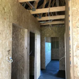 Wnętrze w trakcie budowy: surowe ściany z płyt OSB, drewniana konstrukcja dachu z widocznymi kablami elektrycznymi, wylewka podłogowa, widok przez otwory drzwiowe.