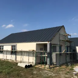 Wznoszony parterowy budynek z ciemną blachodachówką, częściowo otynkowany, z widocznymi rusztowaniami i elementami wykończeniowymi wokół.