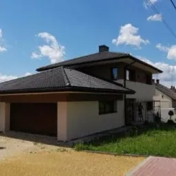 Dwukondygnacyjny dom jednorodzinny z garażem w trakcie prac elewacyjnych, widoczne rusztowania i pracownik przy elewacji, jasna elewacja i ciemna dachówka.