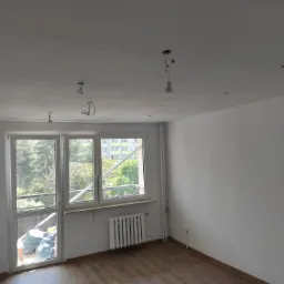 Pok&oacute;j po szpachlowaniu ścian i sufitu, widoczne ślady po instalacji oświetlenia, panele podłogowe, okno z widokiem na zewnątrz, grzejnik i worki z odpadami budowlanymi przy balkonie.