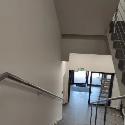 Klatka schodowa w budynku biurowym, świeżo po malowaniu ścian na jednolity, jasny odcień szarości, widoczna metalowa barierka i schody wyłożone płytkami, w tle otwarte drzwi z zielonym znakiem...