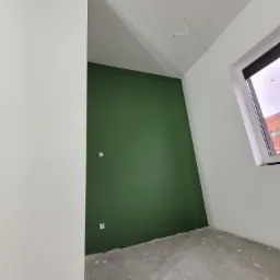 Świeżo pomalowany pokój z jedną ścianą w odcieniu butelkowej zieleni, widoczne gniazdka elektryczne, surowa podłoga przed wykończeniem, białe ściany i okno.