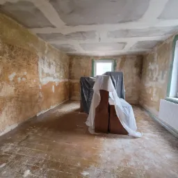 Pokój w trakcie remontu: ściany z resztkami starej farby, częściowo zagruntowany sufit, zabezpieczone meble folią malarską, przygotowanie do gładzi.