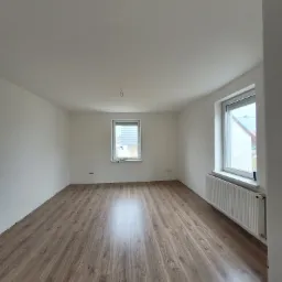 Pusty pokój po remoncie z panelami podłogowymi w odcieniach brązu, świeżo pomalowanymi białymi ścianami i grzejnikiem pod oknem.