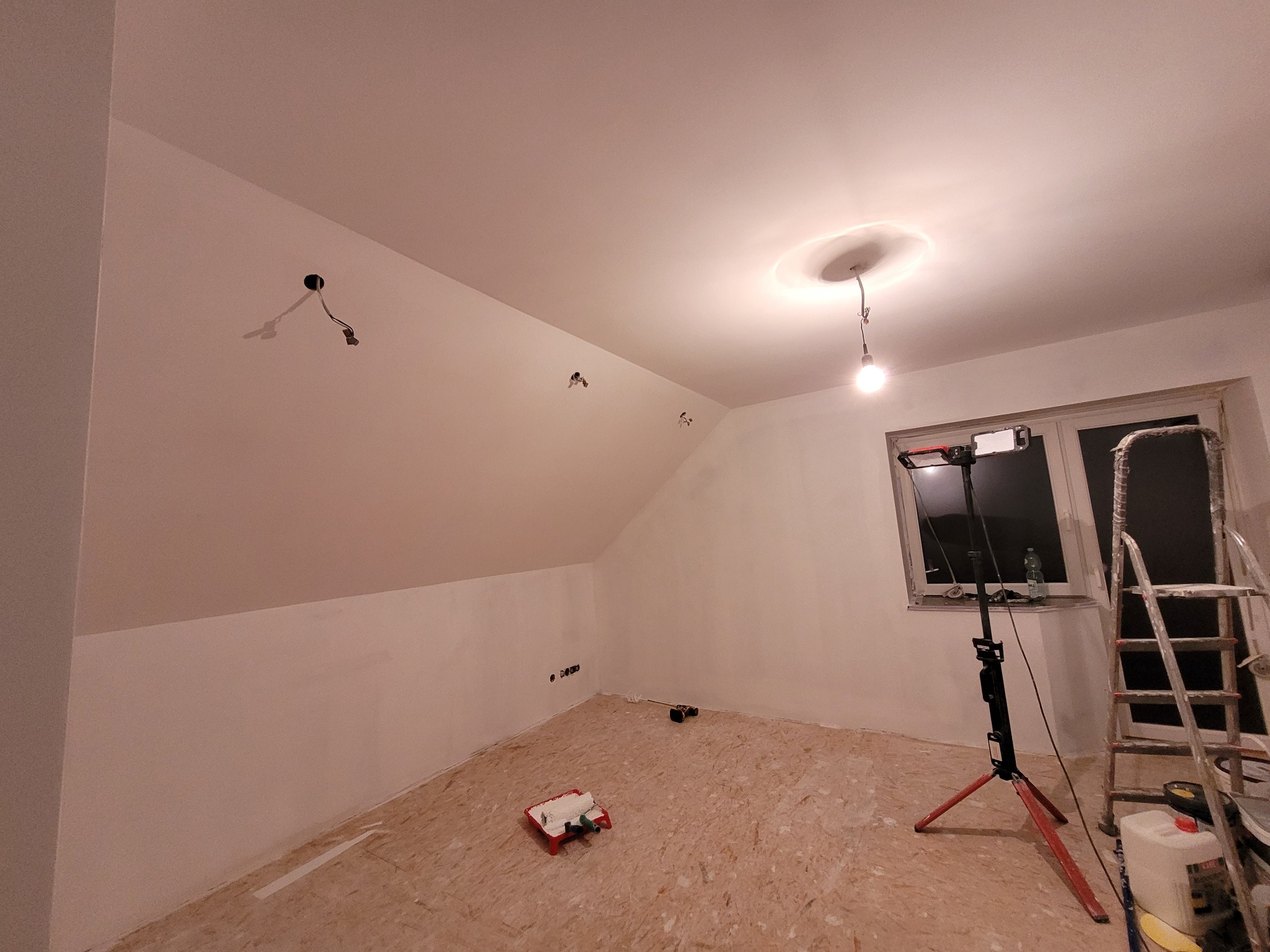 Pokój na poddaszu w trakcie malowania, ściany częściowo pomalowane na biało, widoczny wałek malarski, drabina, lampa na statywie i wystające przewody elektryczne.