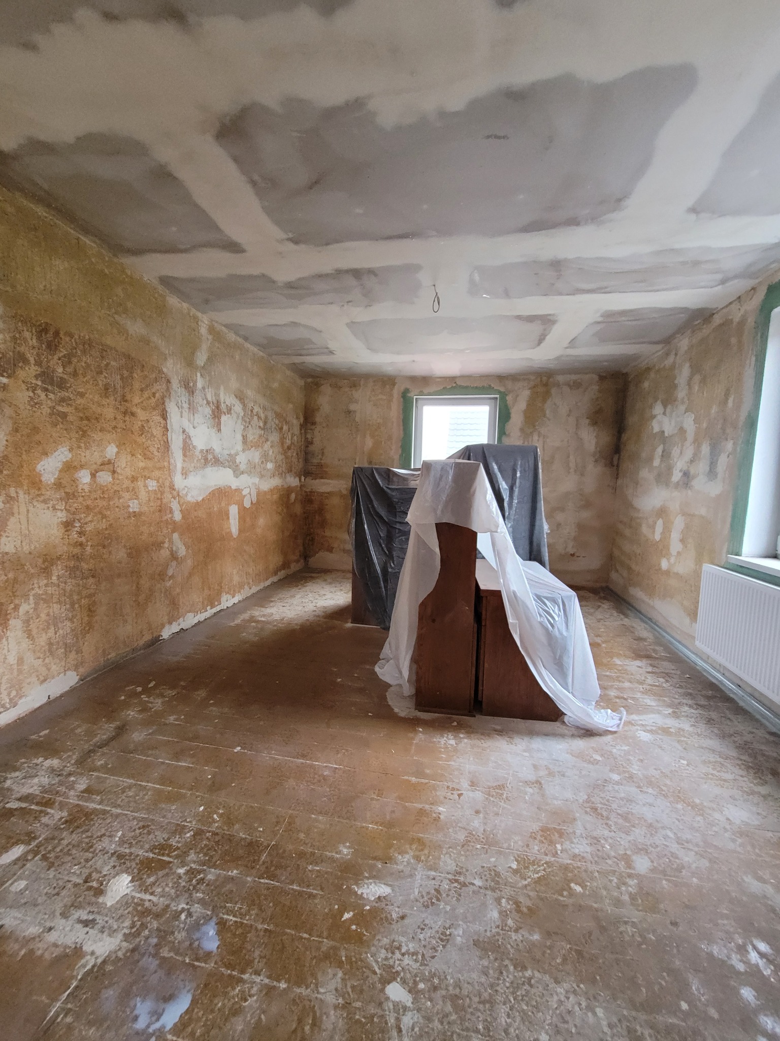 Pokój w trakcie remontu: ściany z resztkami starej farby, częściowo zagruntowany sufit, zabezpieczone meble folią malarską, przygotowanie do gładzi.