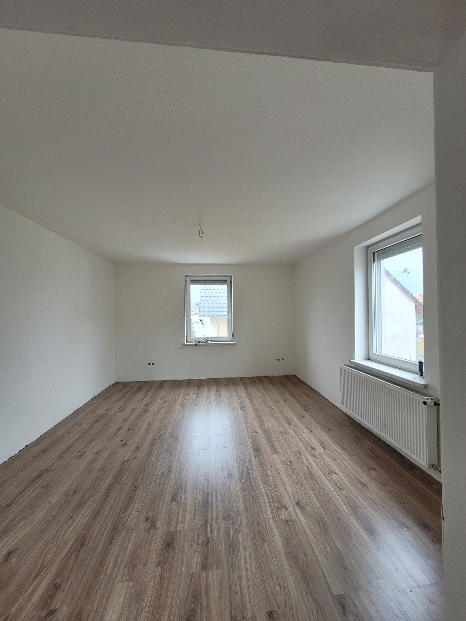 Pusty pokój po remoncie z panelami podłogowymi w odcieniach brązu, świeżo pomalowanymi białymi ścianami i grzejnikiem pod oknem.