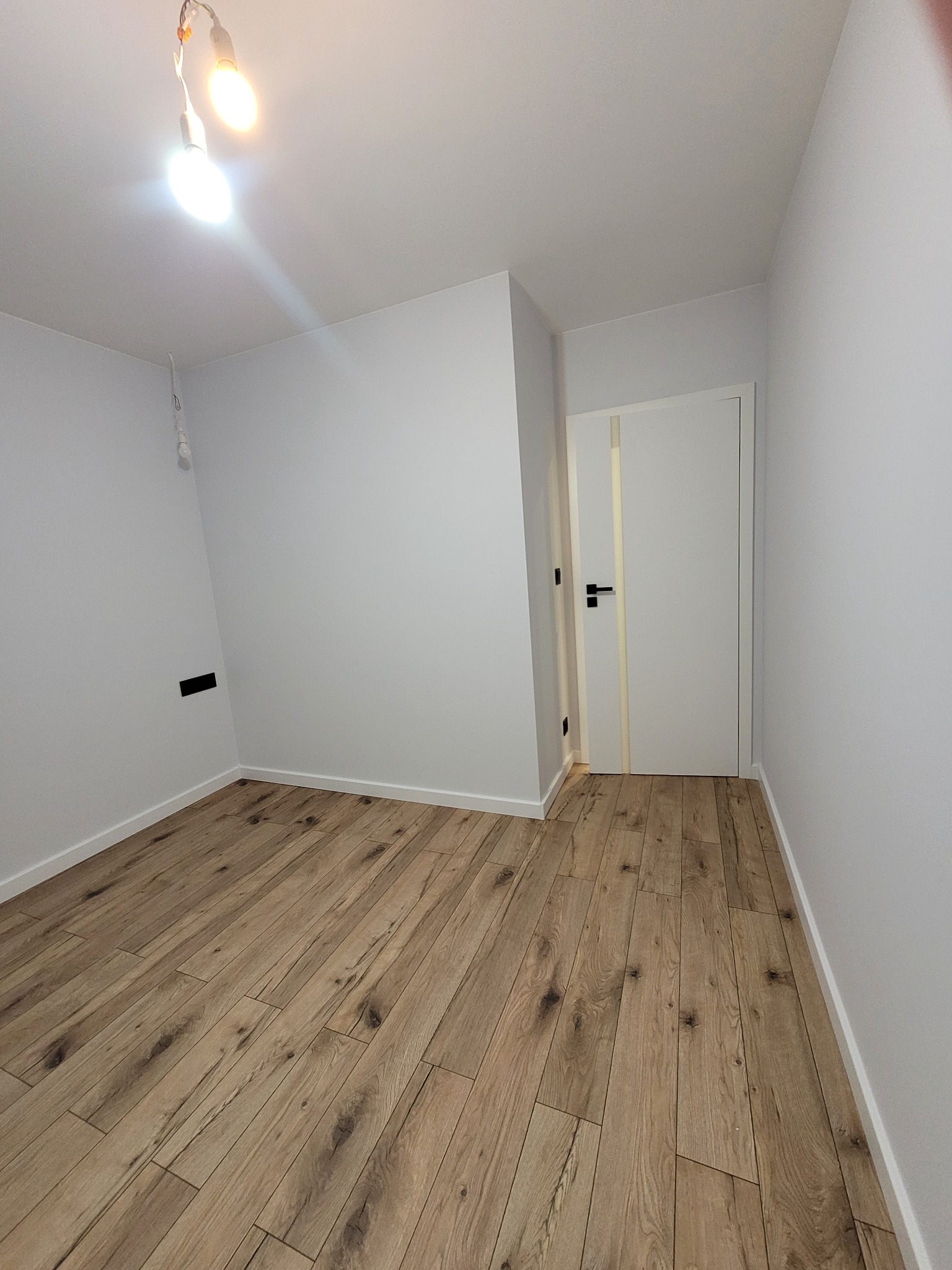 Pusty pokój po remoncie z panelami podłogowymi w odcieniach brązu i beżu, białymi ścianami, białymi drzwiami z pionowym paskiem w kolorze paneli i niedokończonym oświetleniem sufitowym.