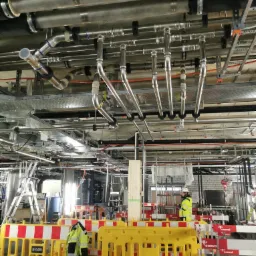 Kompleksowy system rur i instalacji pod stropem w hali przemysłowej, zabezpieczony żółto-czerwoną barierką. Widoczny pracownik w kasku i odblaskowej kurtce.