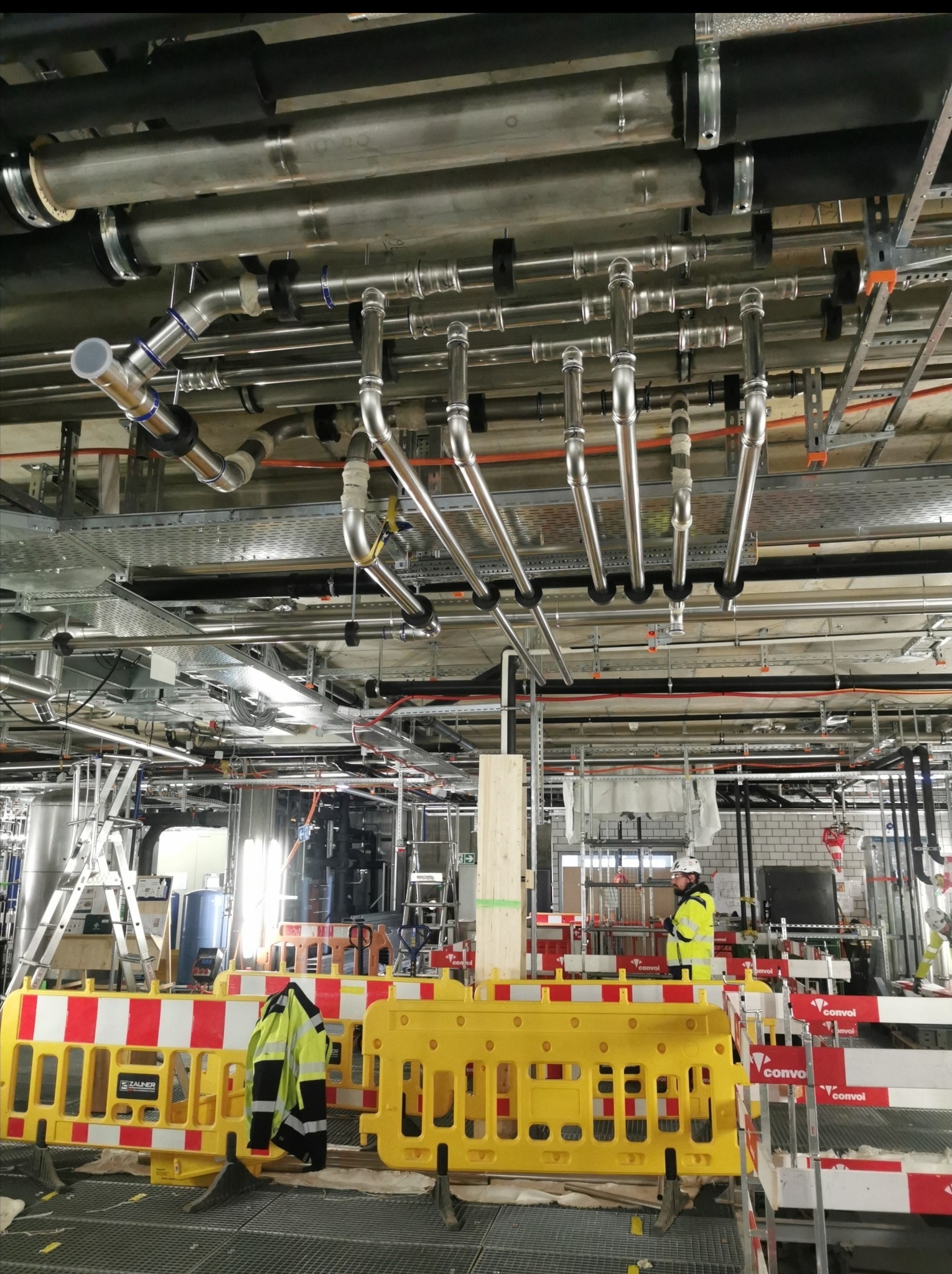 Kompleksowy system rur i instalacji pod stropem w hali przemysłowej, zabezpieczony żółto-czerwoną barierką. Widoczny pracownik w kasku i odblaskowej kurtce.