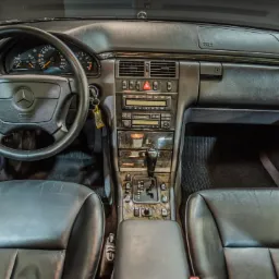 Wnętrze klasycznego Mercedesa W210 z czarną skórzaną tapicerką, widok na deskę rozdzielczą, kierownicę z logo, automatyczną skrzynię biegów i elementy wykończenia z drewna.